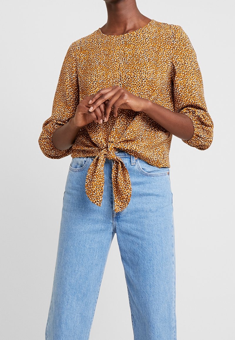 Braune Bluse mit floralem Muster, rundem Ausschnitt, langen Ärmeln und einem gebundenen Vorderteil, kombiniert mit hellblauen hochgeschnittenen Jeans.