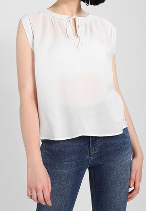 Blusa blanca de manga corta con un escote tipo ojal y lazo, de tejido texturizado y un corte holgado, combinada con jeans de mezclilla azul. Fondo liso.