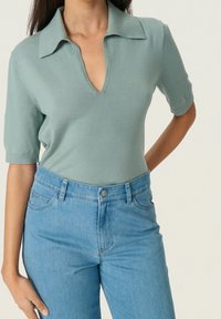 Vaaleanvihreä neulottu yläosa, jossa leveä kaulus ja lyhyet hihat, yhdistettynä korkeavyötäröisiin vaaleansinisiin farkkuihin, joissa edessä napitettava suljenta.