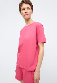Femme aux cheveux courts portant un t-shirt et un short assortis de couleur rose vif, regardant légèrement sur le côté sur un fond uni.