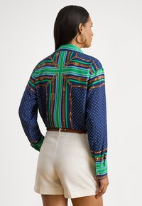 Kvinne med langt mørkt hår som har på seg store hoop-øredobber, en mønstret langermet skjorte med blå, grønne og brune striper, og beige shorts.