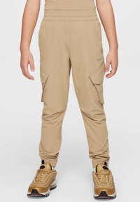 Gyermek bézs cargo nadrágban és arany Nike tornacipőben áll egy sima fehér háttér előtt.