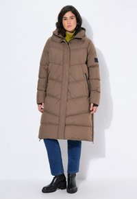Manteau rembourré marron avec capuche, présentant un design matelassé, un zip frontal complet et des poches latérales. Porté par-dessus un col roulé vert et un jean bleu.