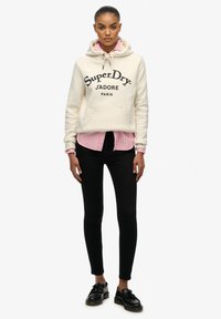 Crème hoodie met de opdruk "SuperDry J'ADORE PARIS", gelaagd over een roze gestreept shirt, zwarte skinny jeans en zwarte schoenen met dikke zolen.