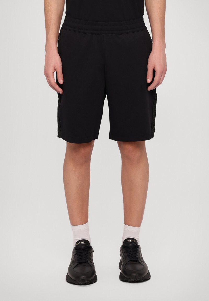 Personne portant un short noir, des chaussettes blanches et des baskets noires, debout devant un fond clair uni.
