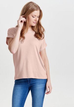 Camiseta de manga corta de color rosa claro, hecha de una tela suave. Presenta un ajuste relajado con mangas enrolladas, combinada con jeans ajustados azules.