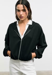Crna zip-up dukserica labavog kroja, s kapuljačom na vezanje i elastičnim manžetama, nosi se preko bijele košulje s kragnom.