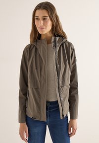 Femme portant une veste à capuche kaki par-dessus un haut beige et un jean bleu, debout devant un fond neutre.