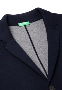 Granatowy kardigan z kołnierzykiem, wykonany z miękkiego materiału, z szarym wewnętrznym wykończeniem i jednym czarnym guzikiem. Widoczna metka marki.