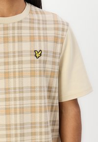 Beige kortermet t-skjorte med rutete mønster i brune og oransje nyanser, med en gul brodert fuglelogo på venstre bryst.