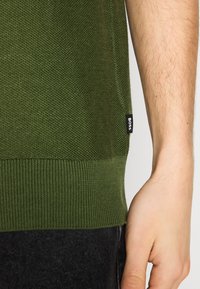 Ourlet de pull texturé vert avec une petite étiquette noire « BOSS » porté par une personne avec un avant-bras et une main nus visibles.