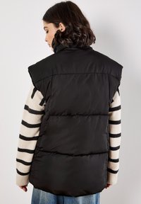 Zwarte gewatteerde vest met hoge kraag, gekenmerkt door brede schoudernaden. Draagt over een gestreepte trui, met een zachte matte textuur.