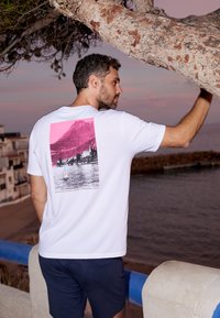 Witte katoenen t-shirt met een roze en zwarte grafiek van een meerlandschap, met bergen en een zeilboot. Draagt met marineblauwe shorts.