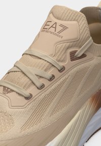 Μπεζ πλεκτό αθλητικό παπούτσι με χοντρή σόλα, μπεζ κορδόνια και λογότυπο "EA7 Emporio Armani" στη γλώσσα του παπουτσιού.