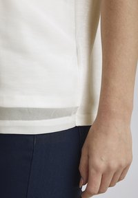 Helles beiges Stoffoberteil mit einer weichen Textur, das über ein unteres Netzpaneel und seitliche Schlitze verfügt. Kombiniert mit dunkelblauen, figurbetonten Hosen.