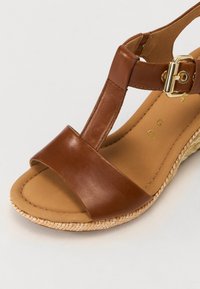 Brown leather wedge sandal with a T-strap design, featuring a gold buckle and a textured, woven jute-like base. Smooth tan insole.