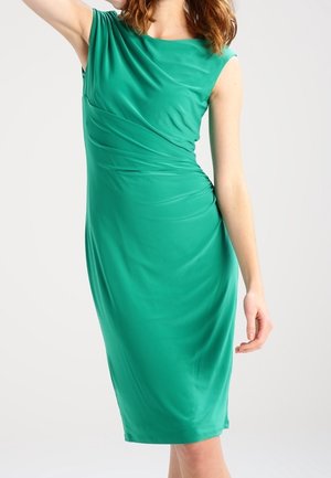 Kvinde iført ærmeløs, knælængde grøn kjole med plisserede detaljer ved taljen, poserende med den ene arm løftet og den anden afslappet.