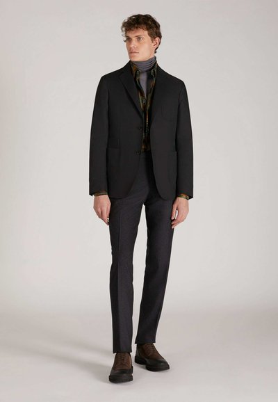 Blazer nero su misura con due bottoni, indossato sopra una camicia a fantasia multicolore e un dolcevita grigio. Pantaloni scuri e testurizzati e scarpe marroni.