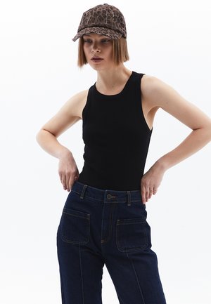 Giovane donna con indosso un top nero senza maniche, jeans blu scuro con grandi tasche frontali e un cappellino a stampa leopardata, in posa con le mani sui fianchi.