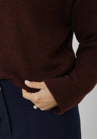 Pull en laine brun avec un ourlet et des poignets côtelés, au fini doux et texturé. La main repose doucement sur l'ourlet. Pantalon foncé visible.