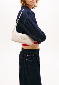 Νεαρό άτομο που φοράει σκούρο τζιν μπουφάν και τζιν παντελόνι, κρατώντας μια μικρή μπεζ τσάντα ώμου με το λογότυπο "tommy jeans".