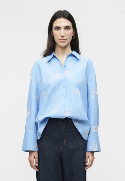 Woman with long dark hair wearing light blue button-up blouse with white floral embroidery and dark high-waisted jeans, standing against light background.