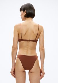 Sieviete ar īsiem tumšiem matiem, ģērbusies brūnā bikini, redzama no aizmugures, stāv pie vienkārša baltā fona.