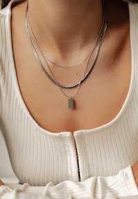 Silberne, mehrlagige Halsketten mit einem flachen rechteckigen Anhänger, einer Schlangenkette und dünnen Ketten, auf einem hellen, gerippten Oberteil.