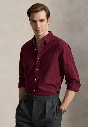 Jeune homme portant une chemise bordeaux à boutons avec manches retroussées et un pantalon plissé gris, debout les mains dans les poches devant un fond neutre.