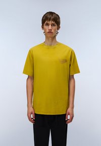 Kortærmet T-shirt i sennepsgul bomuld med rund hals og lille grafisk tryk på venstre bryst. Glat tekstur, afslappet pasform.
