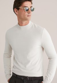 Heled hall, pika varrukatega, kõrge kaelusega kampsun pehmest kangast, koos selge, ümarate prillitega. Tume vöö hõbedase pandlaga.