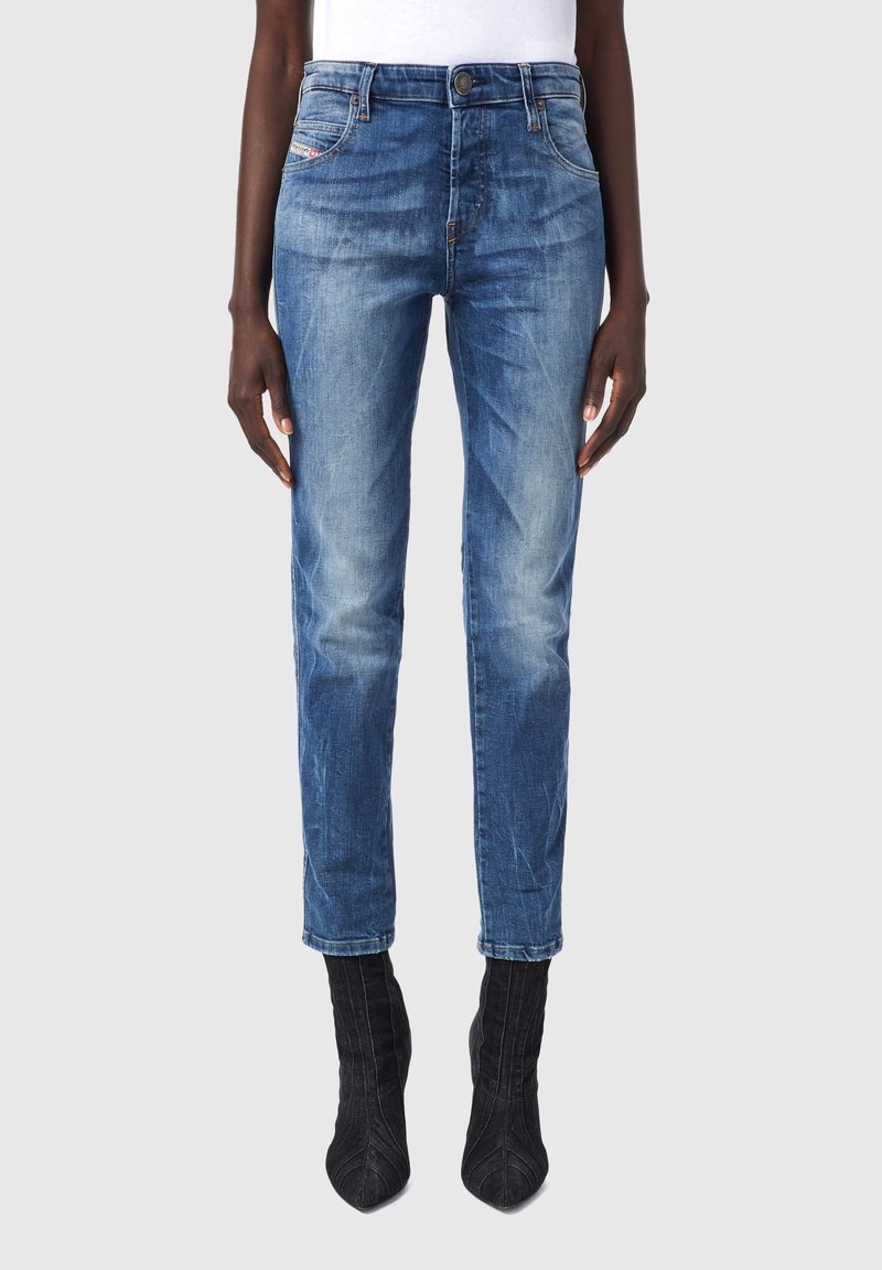 Jean bleu en denim slim fit porté avec des bottines noires à talons et un haut blanc, montré de la taille aux pieds sur une personne debout.