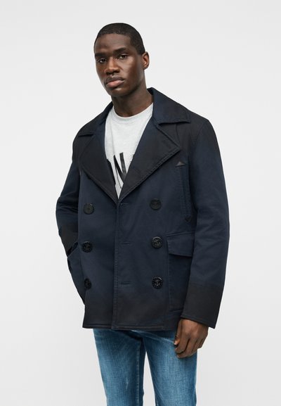 Homme portant un caban bleu marine à double boutonnage sur une chemise gris clair et un jean bleu, debout avec une main dans la poche du manteau.