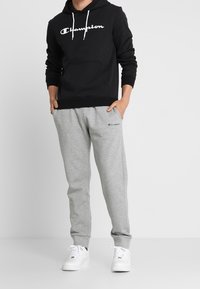 Felpa nera con cappuccio e logo bianco "Champion", abbinata a pantaloni di felpa grigi e sneakers bianche. Il tessuto appare morbido e casual.