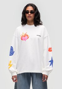 Felpa bianca oversize con grafiche stampate colorate: un cuore rosa con fiamme, stelle e testo sulle maniche lunghe. Design semplice con collo a giro.