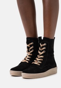Stivali alla caviglia neri in suede con una piattaforma beige testurizzata, dotati di occhielli metallici e spesse lacci intrecciati beige. Chiusura con zip sul lato.