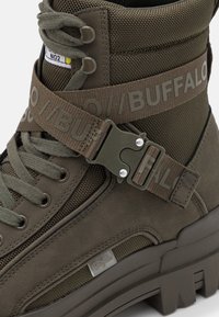 Vysoké olivově zelené boty s texturovanou tkaninou, semišovými detaily, páskem s logem "BUFFALO" a kovovou sponou, silnou podešví a tkaničkami.