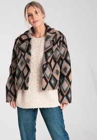 Gekürzte Jacke aus gemustertem Stoff mit Rautenformen in Schwarz, Beige und Hellblau, kombiniert mit einem cremfarbenen Strickpullover und blauen Jeans.