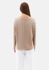 Beige, strukturiertes Langarmoberteil mit weitem Ausschnitt und abgerundetem Saum, von hinten betrachtet. Kombiniert mit weißen Hosen.