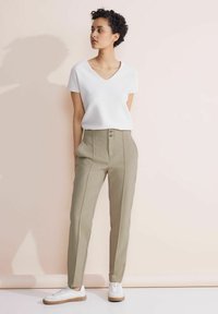 Top bianco a righe con maniche corte, pantaloni beige su misura con cucitura frontale e due bottoni, abbinati a sneakers bianche con suola marrone chiaro.