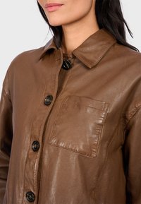 Giacca in pelle marrone con colletto, superficie testurizzata, bottoni neri e taschino sul petto. Presenta una vestibilità comoda e una superficie liscia.