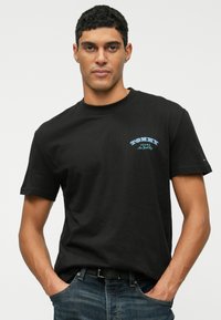 Juodas medvilninis marškinėliai su trumpomis rankovėmis, apvalia iškirpte ir mėlynu grafiniu tekstu ant krūtinės. Kasdieniškas siluetas, derinamas su tamsiais džinsais.