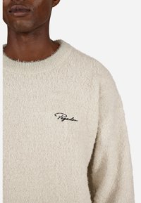 Suéter felpudo de tom bege claro, com decote redondo e tecido texturizado. Apresenta um pequeno logótipo preto bordado na zona superior esquerda do peito.