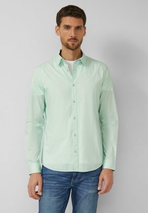 Mann trägt ein hellgrünes Hemd über einem weißen T-Shirt und blauen Jeans, steht vor einem schlichten hellgrauen Hintergrund.