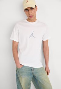Valge puuvillane T-särk, millel on hall Jordan logo rinnal, koos heledate denim teksaste ja heledat värvi kollase mütsiga, millel on punane logo.