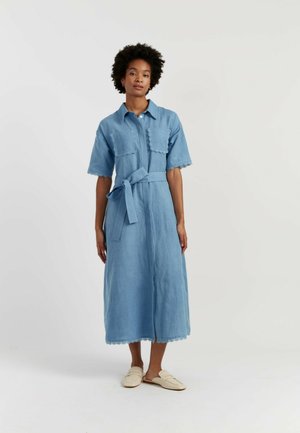Donna in piedi che indossa un vestito midi azzurro chiaro con maniche corte, tasche frontali, cintura in vita e scarpe crema slip-on.