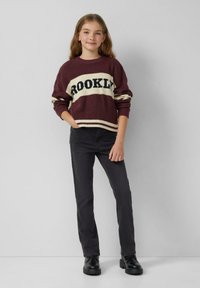 Młoda dziewczyna z długimi włosami, ubrana w bordowy i kremowy sweter z napisem "Brooklyn", czarne dżinsy i czarne botki, stoi z jedną ręką w kieszeni.