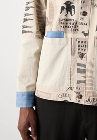 Baumwolljacke mit einem beigen Grundton und schwarzen grafischen Mustern, blauen Akzentärmeln und Vordertaschen. Holzknebel vervollständigen das Design.