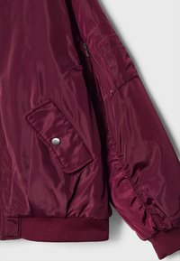Manche d'une veste bomber bordeaux avec poche zippée et poche à rabat à bouton-pression, poignet et ourlet élastiques.