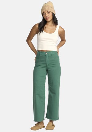 Pantalons larges verts avec des ourlets effilochés, associés à un débardeur blanc ajusté et des sabots en tan, surmontés d'un bonnet en tricot beige.